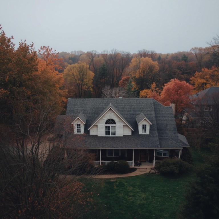 Aerial view of a two-story home with gray asphalt shingle roof, dormer windows, and a wraparound porch, surrounded by trees in full autumn color on an overcast day.