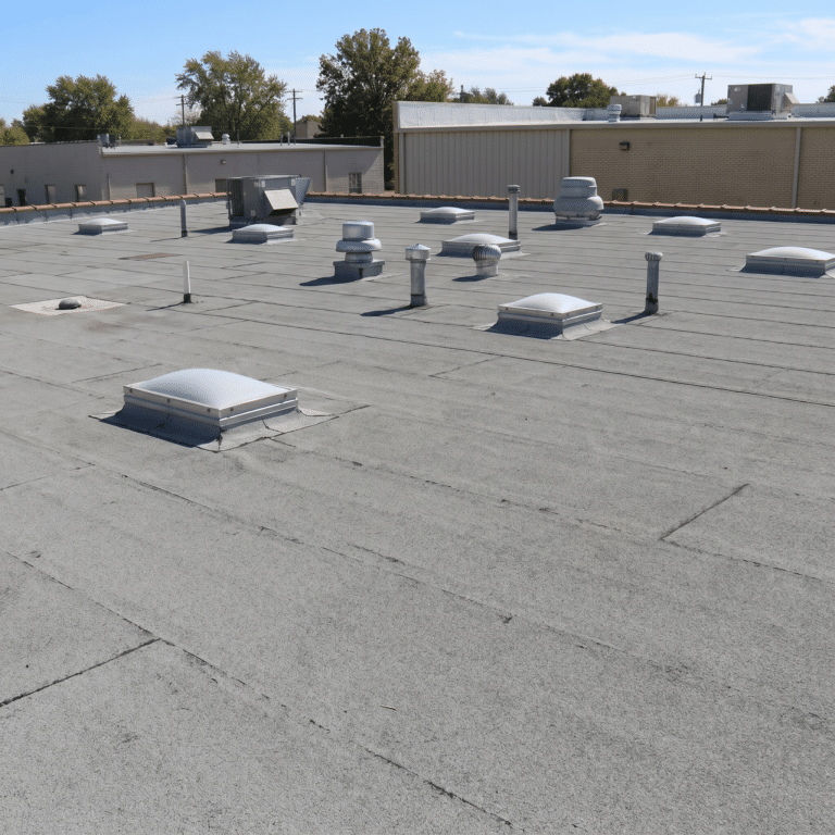 A large flat roof -with a low slope covered in a gray granulated membrane, dotted with pipe vents, turbine vents, and domed roof units, with rooftop equipment and low buildings behind it.
